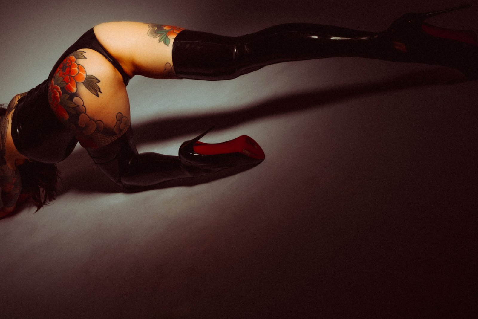 Person with large tattoos wearing black latex thigh-high boots and high heels, posing on hands and knees.