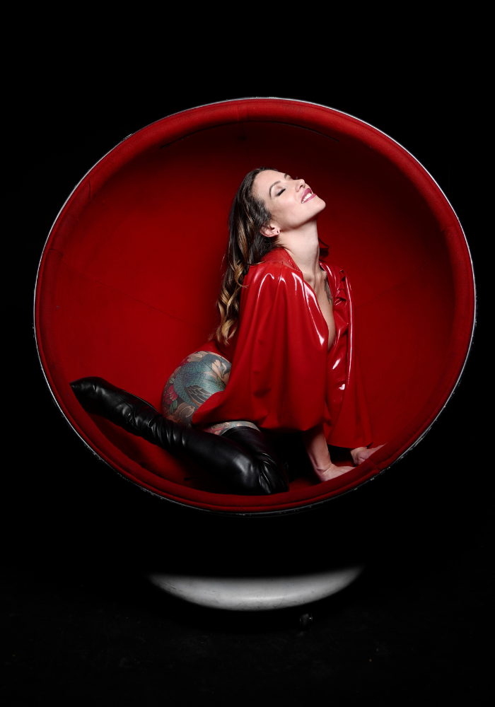 Adreena in a red latex outfit with leather high-healed boots. She's kneeling in a red oval chair, curving her back.