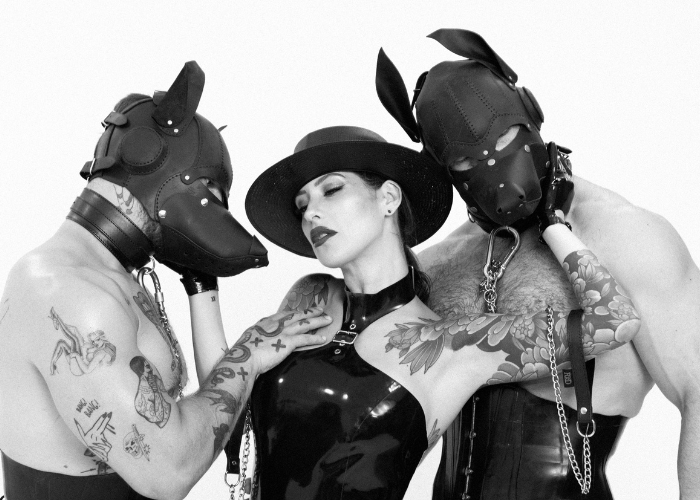 Adreena and two human pups in their masks and leashes. She's holding both of their faces as they touch her back and chest.