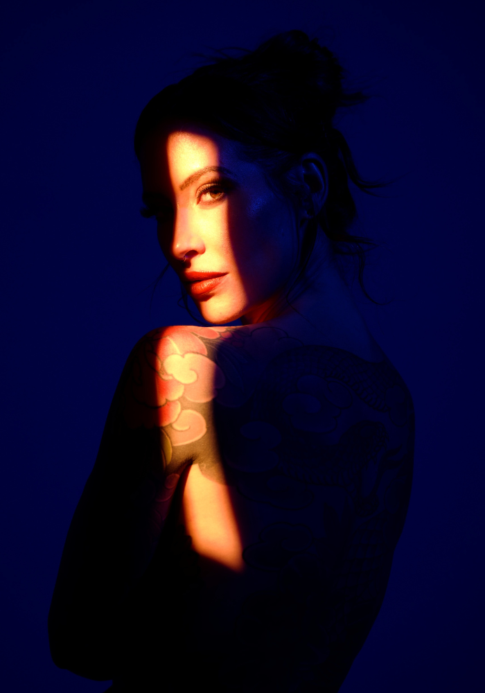 Adreena in a dark blue room, lit with a sliver of golden warm light.