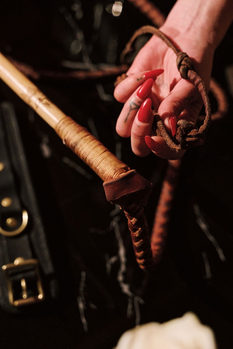Hand with red nails holding a braided whip with a wrapped wooden handle on a dark background.