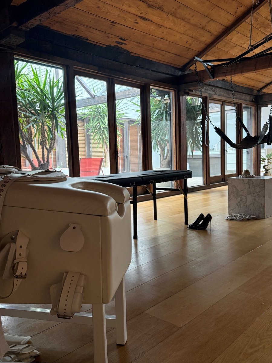 Loft room with wooden floor, large windows, a massage table, hanging swing, and leather straps on a chair.