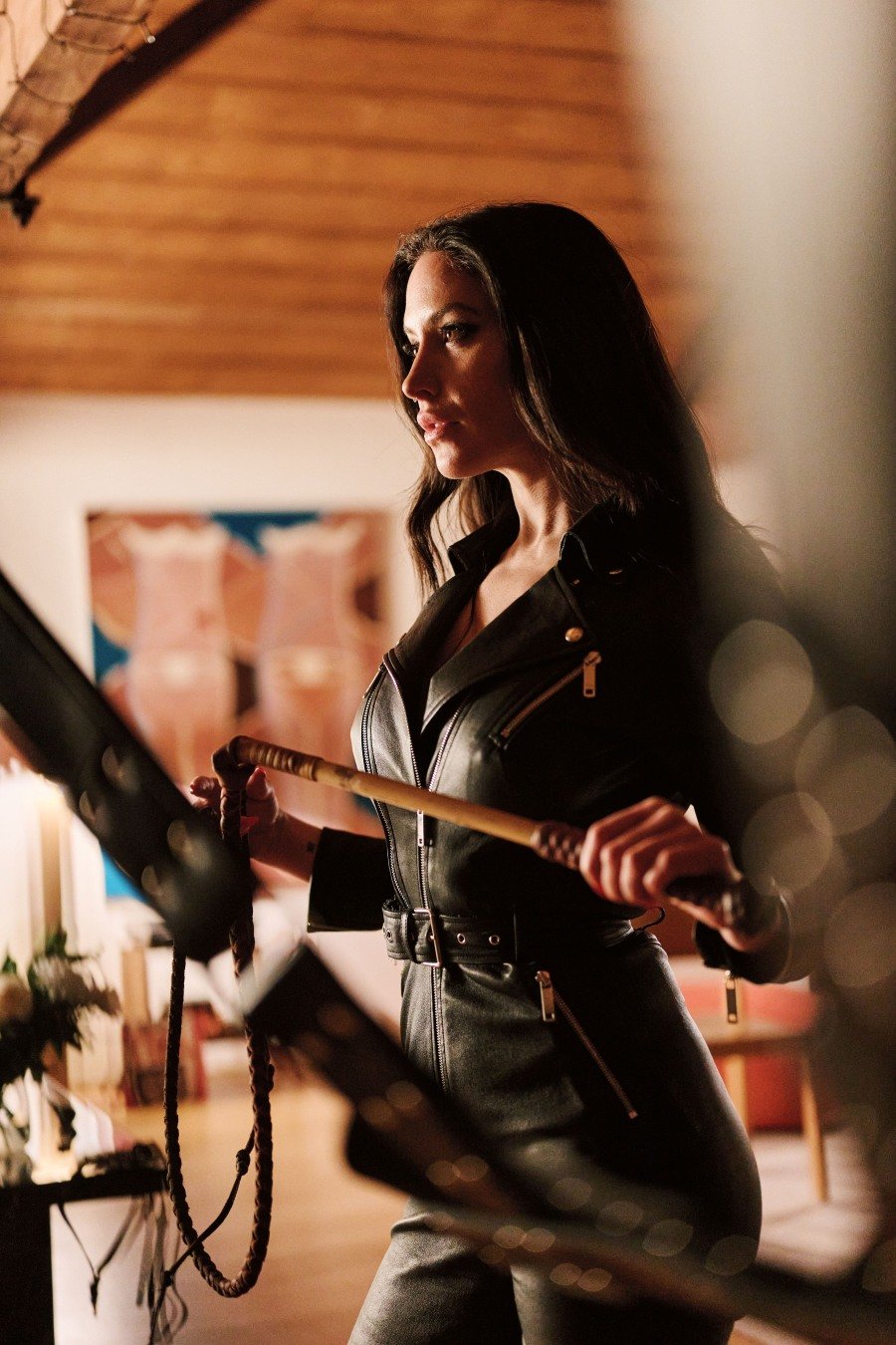 Woman in black leather outfit holding a whip in a warmly lit room with wooden ceiling.