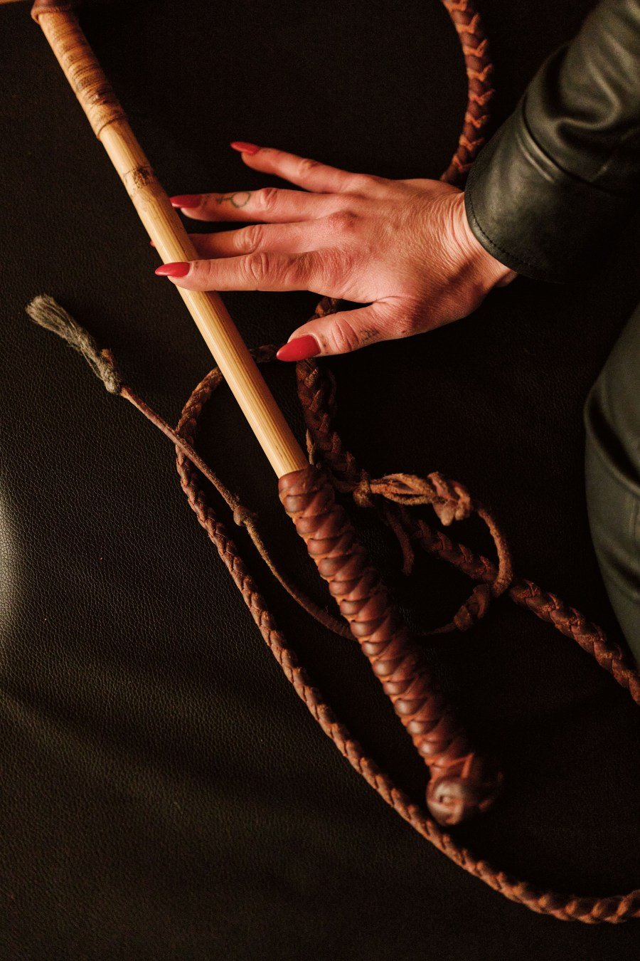 Hand with red nails resting on a braided leather whip with a wooden handle on a dark surface.