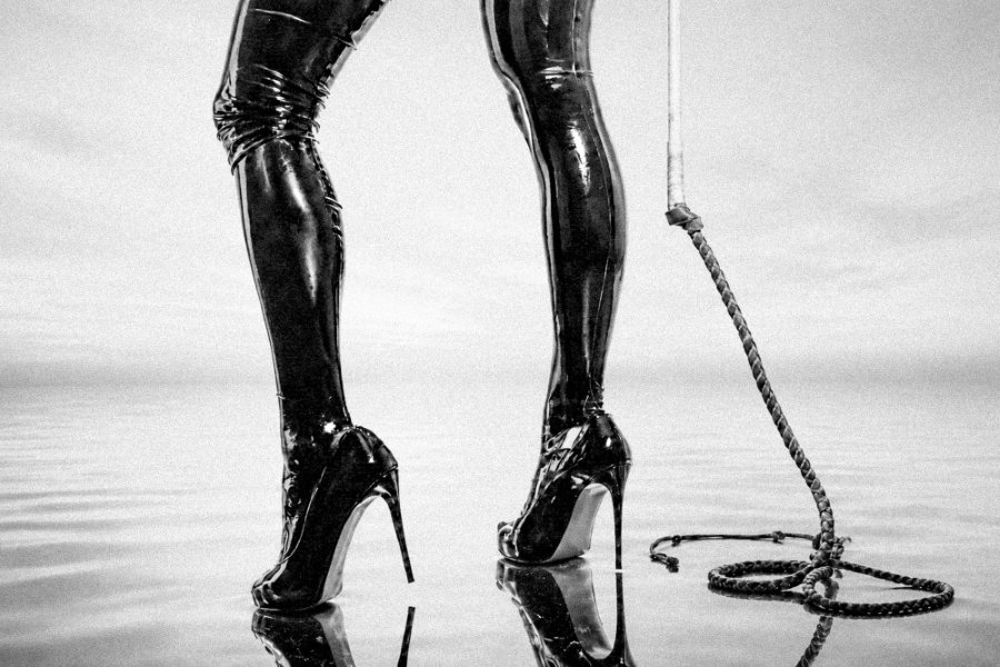 Person wearing shiny black thigh-high boots with heels standing near a coiled rope on reflective surface.