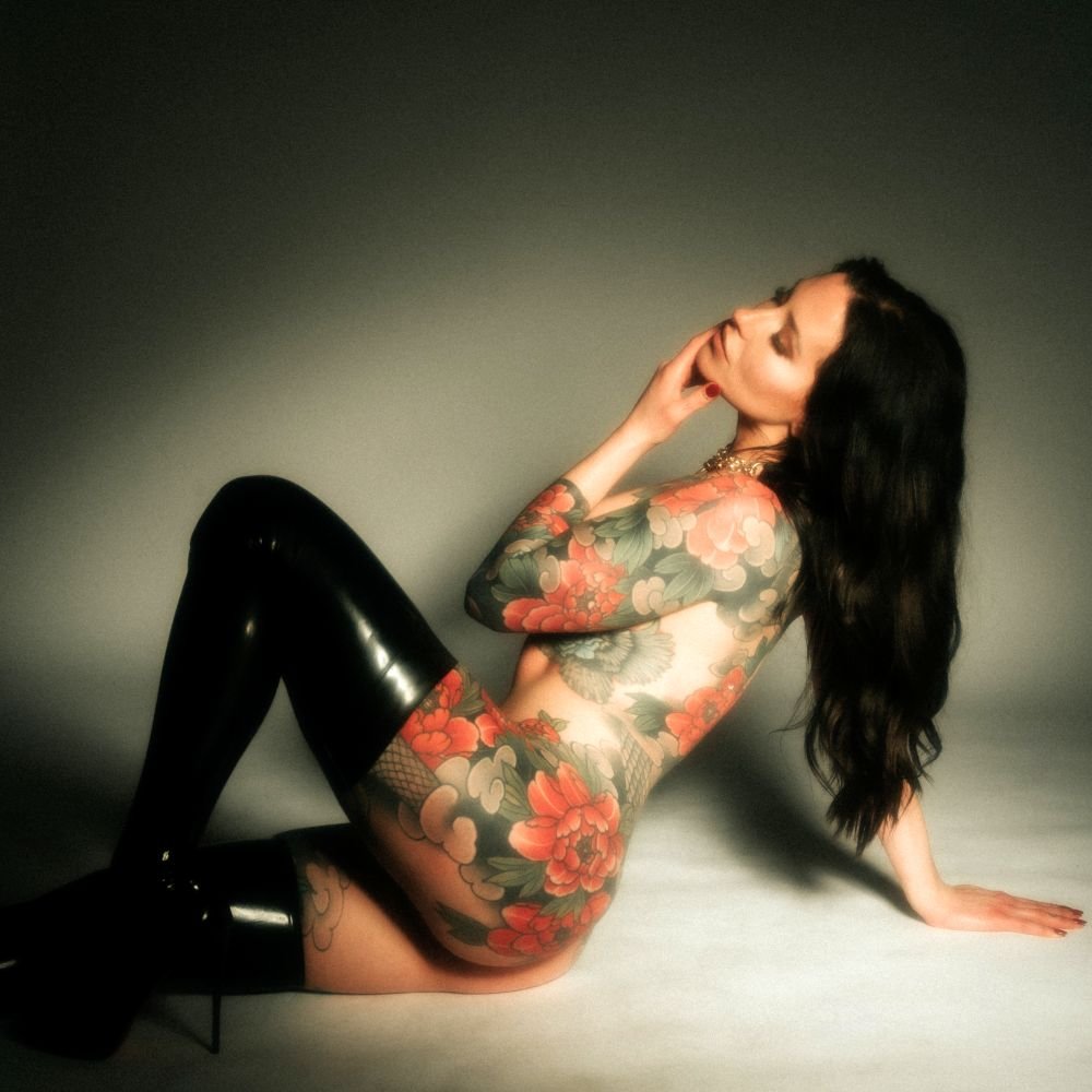 Woman with floral tattoos wearing black thigh-high boots, posed sitting on the floor in side profile.