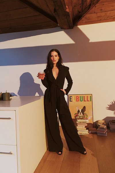 Woman in black suit holding a drink, standing indoors near books and an Art poster on wooden floor.